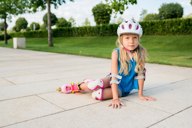 孩子们在公园里学滑旱冰。女孩跌倒后坐在小路上。儿童积极健康的休闲和户外运动。图片下载