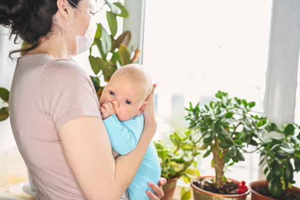 美丽的母亲戴着防护口罩，抱着她可爱的新生儿子，保护他免受病毒和感染。冠状病毒covid-19预防。居家隔离。家人在家靠窗。图片下载