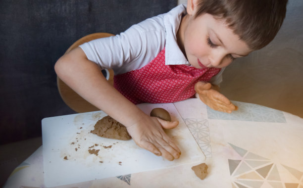 The boy, leaning against the table, rolls a piece of clay. 选择性图像聚焦和声音效果。图片下载