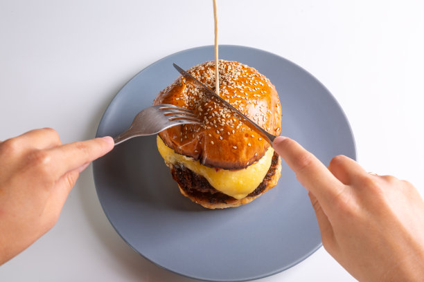 一个男人用刀叉在白色背景的灰色盘子上用刀叉切的自制芝士汉堡。手在画框里。从以上观点。水平方向。高质量的照片图片下载