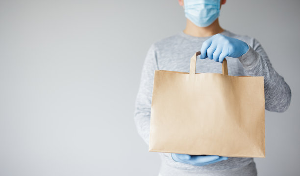冠状病毒和非接触投递——年轻的投递员戴着医用口罩和手套，手持纸袋，背景为白色图片下载