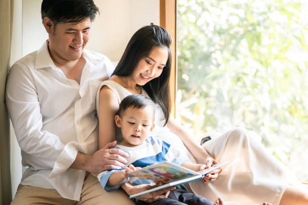 家庭幸福的活动。亚洲母亲拿着书给坐在沙发上的儿子。可爱的小朋友在书上画画。英俊的爸爸和漂亮的妈妈看着它。图片下载