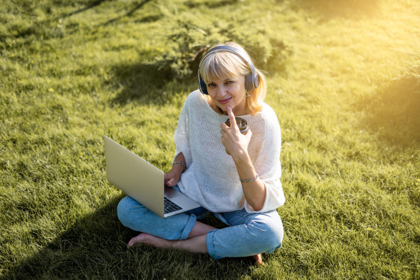 一个成年女子在公园外面的草地上戴着耳机发短信。工作和喝咖啡。使用电脑。远程学习、在线教育、工作和音乐的概念图片下载