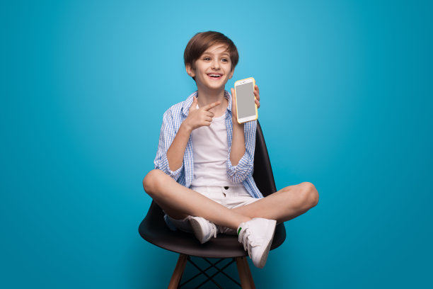 一个快乐的白人男孩拿着手机坐在椅子上，指着工作室蓝色墙壁上的屏幕图片下载