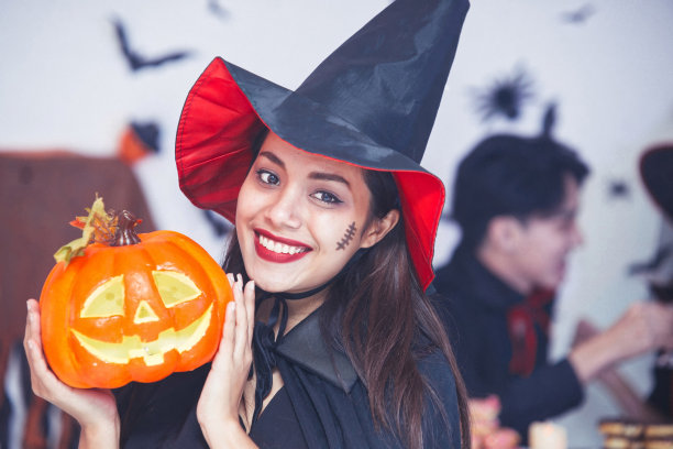 万圣节巫婆拿着南瓜灯。一个迷人的年轻女子的肖像戴着女巫的帽子和美丽的衣服微笑在相机。万圣节晚会背景。图片下载