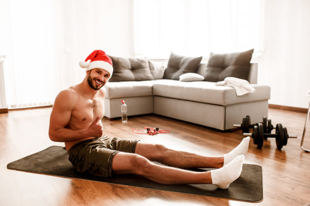 圣诞快乐，恭贺新禧!戴着圣诞红帽的家伙在家里锻炼。他坐在一块裸露上身的地毯上，竖起大拇指。图片下载