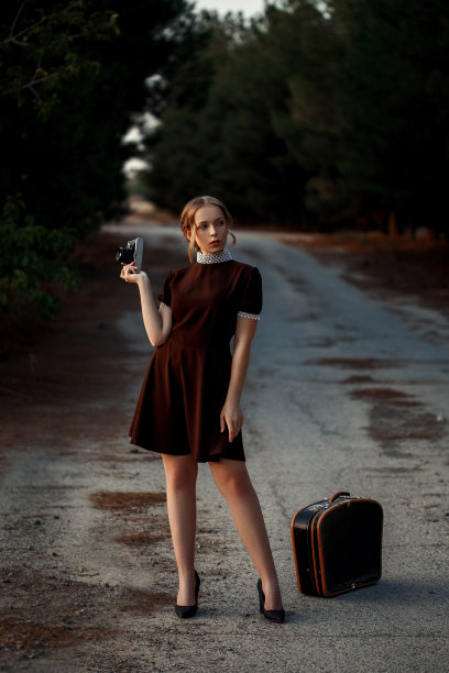 在一条废弃的路上，一个年轻漂亮的女孩穿着一件棕色复古裙，手里拿着一架老式相机，旁边是一个黑色的行李箱图片下载