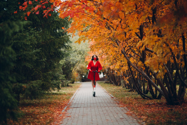 穿着红色连衣裙的年轻女子骑着电动摩托车在秋天的城市公园里图片下载