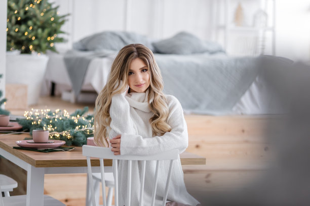 美丽的金发卷发和长头发的圣诞树旁的灯的花环。一个女孩的特写肖像，带着白色的微笑，穿着灰色的针织毛衣，领子很大。复制空间，纹理图片下载