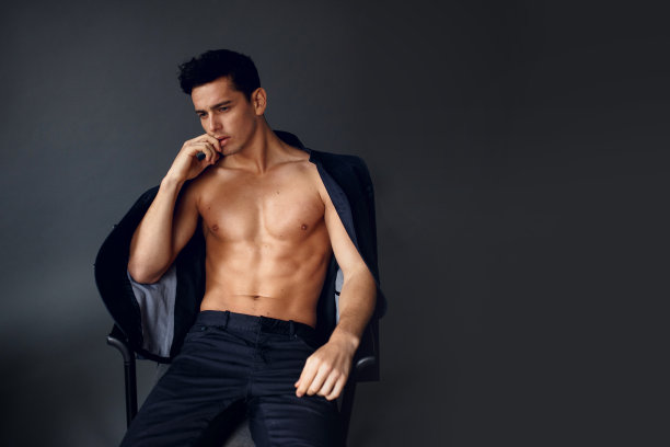 一个优雅的赤膊的黑发男子，肩上披着夹克，坐在椅子上，背景是灰色的。图片下载