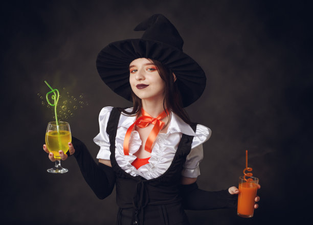 万圣节派对。美丽的女孩在黑帽子和巫婆服装持有魔术鸡尾酒在她的手在黑色雾背景图片下载