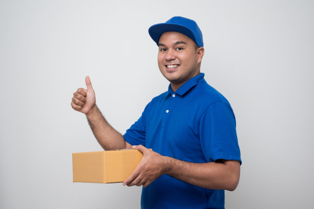 年轻微笑的亚洲快递员在蓝色制服拿着盒子包裹纸板和显示大拇指在孤立的白色背景。图片下载