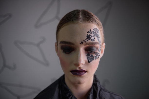 一个女孩的特写，干净的皮肤，明亮的专业化妆，紫色的口红，脸上的黑色常春藤纹身在她的眼睛周围的莱茵石。灰色背景，衣架图案。时尚图片下载