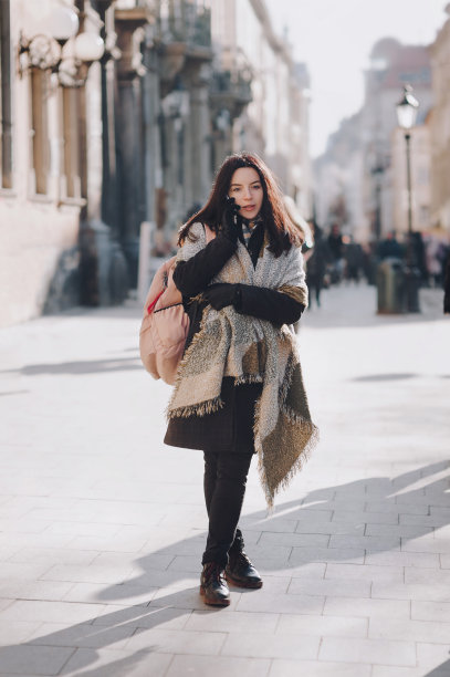 一个黑头发的小女孩，穿着一件深色羊毛外套，裹着宽围巾，穿着靴子，站在阳光普照的街道中央，专心地讲着电话。利沃夫,乌克兰。图片下载