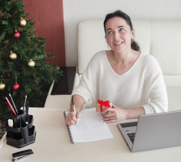 年轻女子在家里的办公室写纸条，手里拿着礼物盒，准备圣诞节。冬天假日销售图片下载