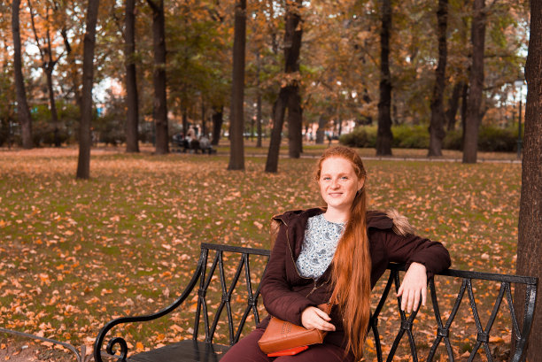 一个留着红色长发的年轻女子坐在公园的长凳上。图片下载