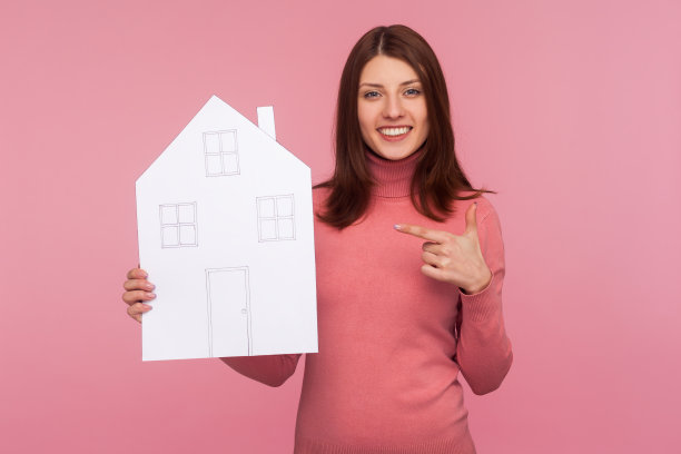 快乐微笑的女人棕色头发在粉红色毛衣指着手指在她的手，看着相机，建筑机构图片下载