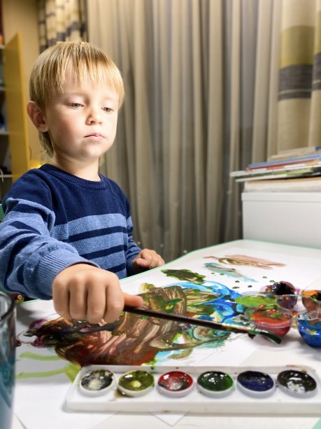一个皮肤白皙的小孩在儿童房的桌子上用多种颜色的颜料画画。图片下载