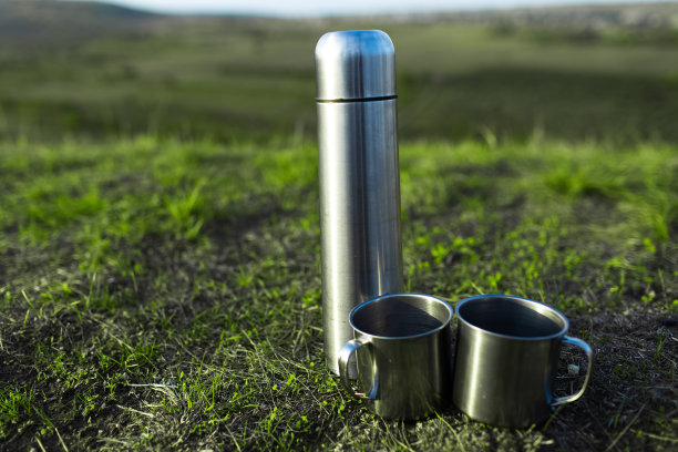 特写的不锈钢热水瓶和绿色草地上的两个杯子。图片下载