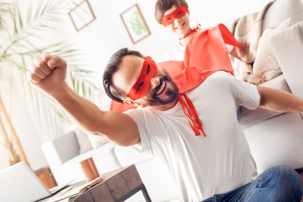 父亲和儿子在超级英雄的服装在家里的地板上的男人手向上飞好玩的特写而男孩坐在沙发后面图片下载