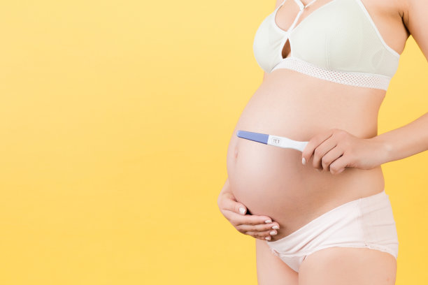 裁剪图像孕妇在彩色内衣显示阳性怀孕测试对她的腹部在黄色背景。分娩的预期。本空间图片下载