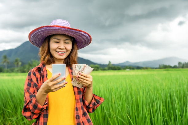 手持泰国钞票和智能手机，站在绿色稻田上的年轻亚洲女农民图片下载