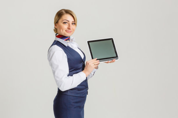 年轻女子专业空姐孤立地站在墙上拿着数字平板电脑显示屏幕看着相机友好地微笑图片下载