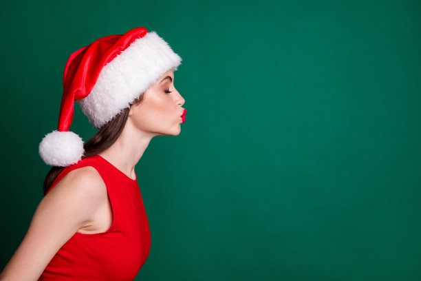 个人资料照片迷人迷人的女士送空中吻空空间魔术奇迹气氛圣诞日期闭上眼睛戴圣诞老人帽红裙子雪女孩派对孤立的绿色背景图片下载