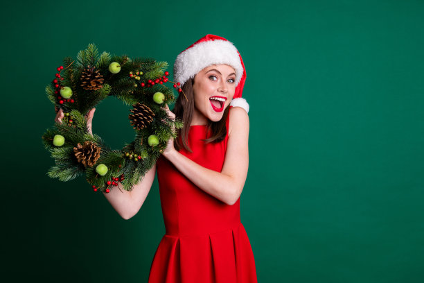 照片惊奇的女孩在圣诞老人帽享受装饰房子在圣诞派对上举行copyspace常绿松树花环穿裙子孤立在绿色背景图片下载