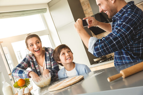 一家人一起站在厨房的桌子上，爸爸在拍妈妈和儿子做面团的照片，笑得很开心图片下载