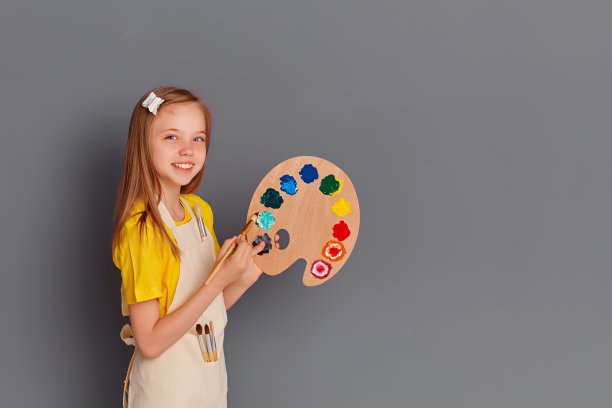 一个漂亮快乐的女孩拿着调色板，用画笔擦着一堵空荡荡的灰色墙壁。她会写问候。空间的文本。本空间图片下载