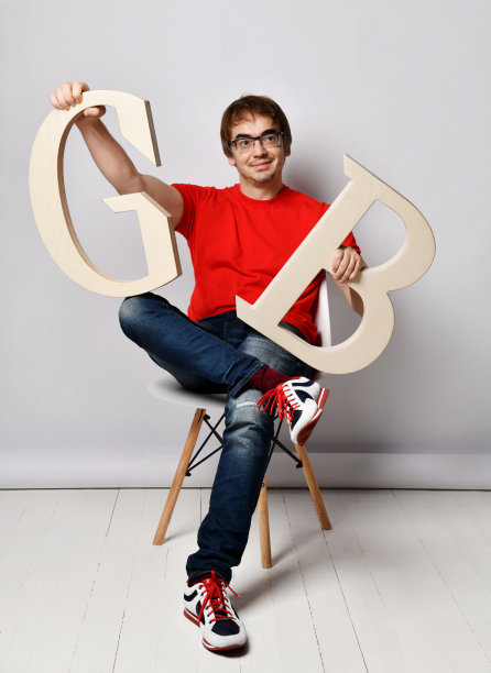 笑容满面的中年男子穿着红色t恤、蓝色牛仔裤、运动鞋和眼镜坐在那里，膝盖上放着大大的字母G和B图片下载