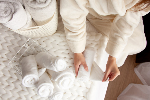 身着米色暖袍的妇女正在折叠干净的白毛巾，并将其放入金属网篮中图片下载