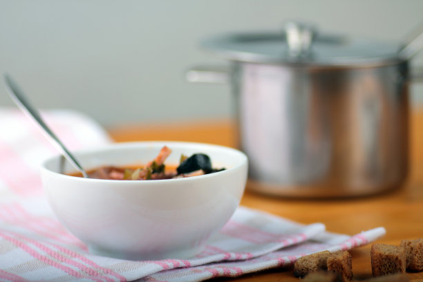 白色的碗里有蔬菜和肉的浓汤，面包丁和平底锅，铺在白色的餐巾上，背景是白色的木头图片下载