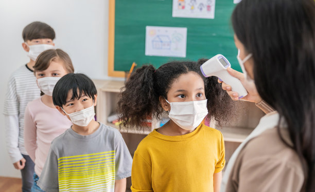 新冠肺炎疫情暴发时，教室里戴医用口罩的黑人女学生肖像。在幼儿园入口处，老师和孩子们拿着温度计。社交距离，冠状病毒预防。学校体温检查。图片下载