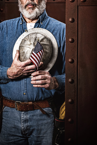 一名男性建筑工人举着一面小美国国旗，手拿安全帽放在胸前图片下载