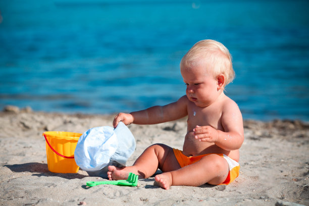 蹒跚学步的孩子坐在沙滩上，在沙子里玩塑料玩具。图片下载