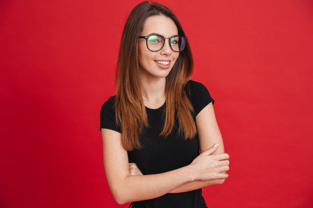 特写照片的现代女性20多岁穿着黑色t恤和眼镜摆姿势与双手折叠和美妙的微笑孤立在红色的背景图片下载