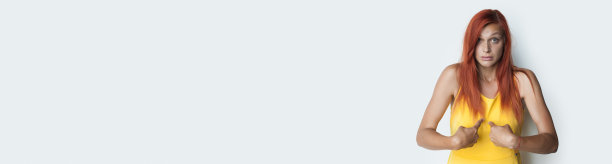 红发女子惊恐地指着她，她穿着一件黄色的t恤，贴在工作室灰色的墙上，墙上有自由空间宣传什么东西图片下载