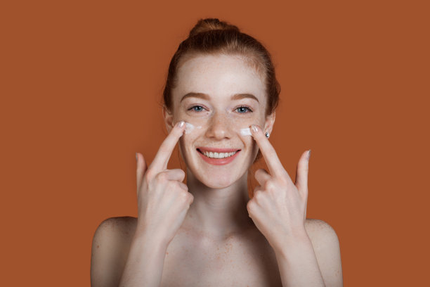 近距离的照片与雀斑姜女士涂抹霜在她的脸上摆姿势与赤裸的肩膀在一个棕色工作室的墙壁微笑的相机图片下载