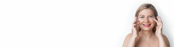 资深金发白种人妇女在她的眼睛附近涂抹抗衰老霜，并对着镜头微笑，在工作室的白色墙壁上有自由空间图片下载