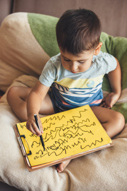 一个白人黑发男孩坐在家里的床上，用马克笔在黄色的纸上画画图片下载