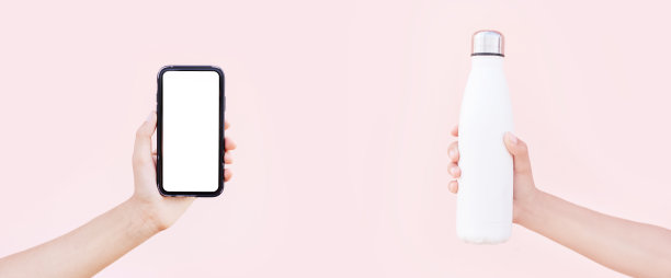 另一只手拿着带模型的智能手机和可重复使用的白色钢制热水瓶。淡粉色的背景。全景概念与复制空间。图片下载