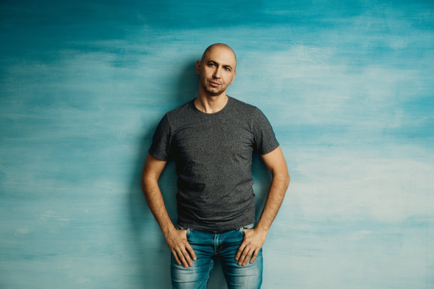 一个穿着灰色t恤的酷酷的秃顶男人在蓝色墙壁的背景下把手插在口袋里摆姿势图片下载