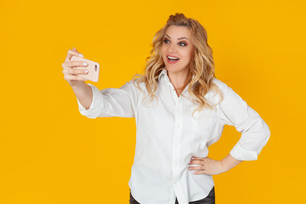 金发女人白衬衫中年欧洲女孩用智能手机自拍，伸出手拿着手机，试图在照片黄色背景下看起来不错的网上生活理念。图片下载