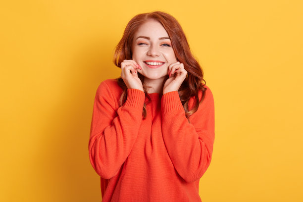 微笑快乐美丽的女士捏她的脸颊，看着微笑直接对着相机和眨眼，穿着橙色毛衣，站在黄色的背景。图片下载