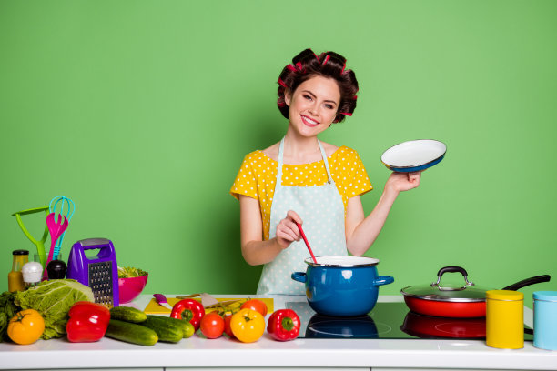 照片积极的女孩准备美味的生态美食晚餐开盖炖锅穿卷发黄点t恤有番茄黄瓜辣椒孤立的绿色背景图片下载