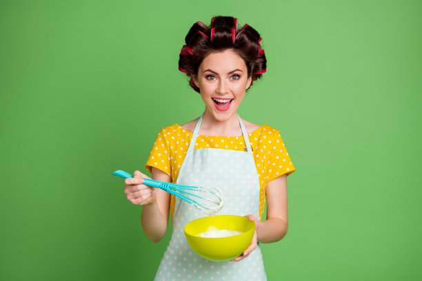 可爱迷人的年轻女孩的照片，滚轴发型，拿塑料打碗，搅拌面团，准备煎饼，家庭午餐，早上，穿有斑点的围裙，衬衫孤立的绿色背景图片下载