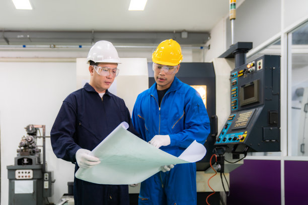 亚洲的工业工程师和工人戴着安全帽讨论蓝图中的产品线，并做出手势和工作在一个重工业制造工厂。图片下载