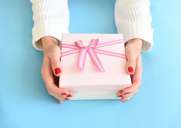 粉色礼品盒，女性手拿蝴蝶结，白色毛衣，红色指甲油，蓝色背景。图片下载